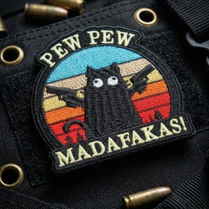 May include: Embroidered patch featuring a black cat holding two guns, with the words "PEW PEW MADAFKASI" above and below the cat. The patch has a retro sunset design with blue, orange, and red colors. The patch is on a black tactical vest.