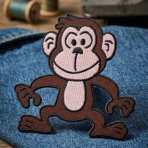 May include: Embroidered monkey patch in brown and pink, with a smiling face and outstretched arms and legs. The patch is on a blue denim fabric, with spools of thread in the background. This patch is suitable for clothing and accessories.