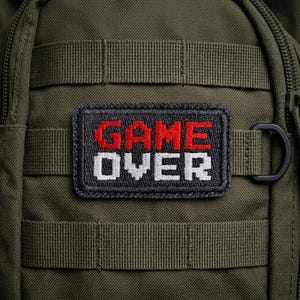 May include: Close-up of an olive green backpack with a black patch. The patch features the words "GAME OVER" in red and white embroidered letters. The backpack has multiple straps and a black D-ring.