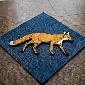May include: Embroidered patch featuring a fox in shades of orange, brown, and white. The fox is depicted walking on a square of blue denim fabric. The patch is placed on a wooden surface.