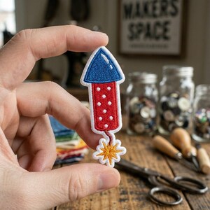 May include: An embroidered patch shaped like a firework rocket with a blue conical top, a red cylindrical body featuring white polka dots, and a yellow starburst explosion at the bottom, all outlined in white thread against a fabric backing.