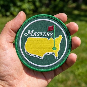 May include: A circular embroidered patch featuring a dark green background with a white circular border. In the center, a yellow map of the United States is displayed with a red golf flag pin marker. The word Masters is stitched in white script.