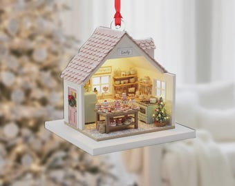May include: A miniature Christmas ornament shaped like a house, featuring a detailed kitchen scene with a table of cakes and a small Christmas tree. The roof is pink, and the house has a white base. The name "Emily" is written on the roof.