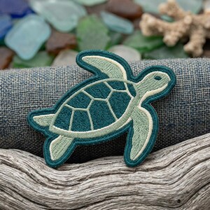 May include: Embroidered sea turtle patch in teal and light green with a cream-colored outline. The patch is on a textured blue fabric, with driftwood and sea glass in the background. The turtle design features a detailed shell pattern.