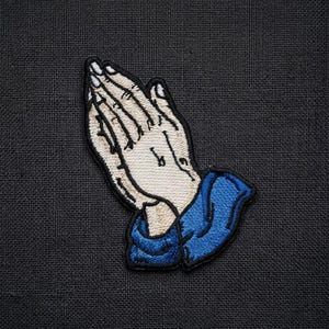 May include: Embroidered patch featuring hands clasped in prayer. The hands are beige with black outlines, and the sleeves are blue. The patch is on a dark gray textured background. This patch is suitable for clothing or accessories.