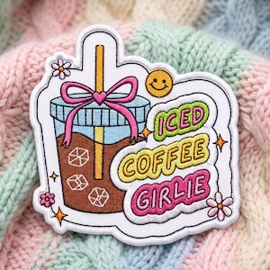 May include: An embroidered patch featuring a brown iced coffee in a clear cup with a yellow straw, a pink bow, and ice cubes. The text reads ICED COFFEE GIRLIE in green, yellow, and pink bubbles. It includes a yellow smiley face and small pink flowers.