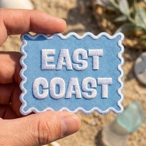 May include: A light blue rectangular patch with a scalloped white border. The patch features the white embroidered text "EAST COAST" centered in a bold, sans-serif font. The design has a clean, stitched finish on a light blue fabric base.