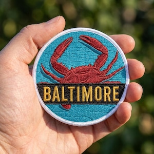 May include: A circular embroidered patch featuring a red crab centered on a teal background. A black horizontal bar across the lower portion contains the word BALTIMORE in bold yellow capital letters. The patch has a white stitched border.