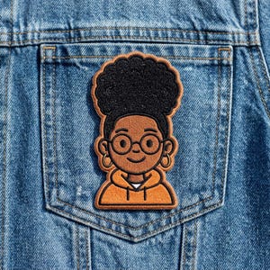 May include: An embroidered patch featuring a cartoon illustration of a person with black afro hair, glasses, and hoop earrings. The person is wearing an orange hoodie. The patch is set against a denim background.