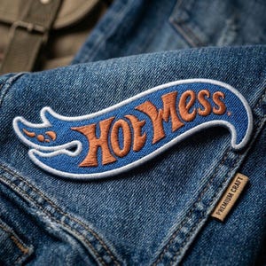 May include: Embroidered patch with the text "Hot Mess" in orange script on a blue background, outlined in white. The patch is sewn onto a denim jacket. A small tag reads "Premium Craft".