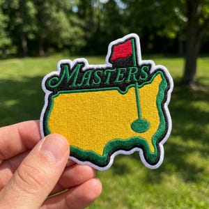 May include: An embroidered patch shaped like the United States map in yellow with a green border and white trim. The word Masters is stitched in green cursive script across the top. A golf flag with a red flag and green pole is visible.
