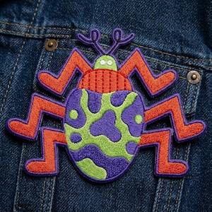 May include: Embroidered patch featuring a colorful bug design. The bug has a purple and green spotted body, orange legs, and purple antennae. The patch is attached to a denim jacket, showcasing its potential use as a decorative element for clothing or accessories.