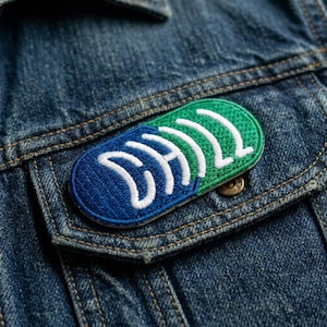 May include: Embroidered patch with a blue and green design, featuring white text. The patch is oval-shaped and sewn onto a denim jacket pocket. The design is abstract and modern, with a clean, minimalist aesthetic.