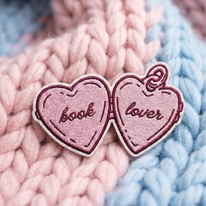 May include: A heart-shaped embroidered patch featuring two connected halves in shades of pink and maroon. The left side displays the word book in cursive script, while the right side displays the word lover in cursive script with a small loop at the top.