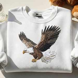 May include: White sweatshirt featuring a detailed embroidered design of a bald eagle in flight. The eagle has brown and white feathers, a yellow beak, and is set against a plain white background. The sweatshirt has a crew neck and long sleeves.