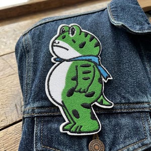 May include: Embroidered patch featuring a cartoon frog with a blue scarf. The frog is green with black spots and a white belly. The patch is on a denim jacket.