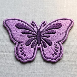 May include: A purple butterfly patch with embroidered details. The patch features a detailed design with darker purple accents on the wings and body. The butterfly is centered on a light gray background.