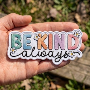 May include: An embroidered patch with a white background featuring the text BE KIND in colorful block letters and the word always in black cursive script. Small white flowers with yellow centers are placed around the lettering.