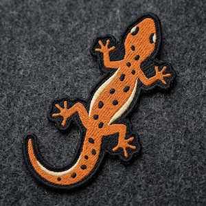May include: Embroidered patch of an orange lizard with black spots and a black outline. The lizard has a white belly and is set against a dark gray textured background. The patch is likely for clothing or accessories.