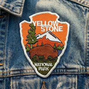 May include: An embroidered arrowhead-shaped patch featuring an orange background with white text that reads YELLOW STONE NATIONAL PARK. The design includes a green tree, a white mountain peak, and a brown bison standing on a green grassy field.