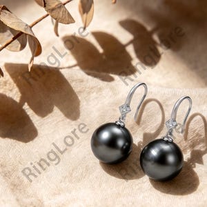 May include: A pair of silver drop earrings featuring round, dark gray pearls suspended from a delicate silver hook. Each earring includes a small, clear, square-cut stone set in a silver mount above the pearl, creating a refined and elegant appearance.
