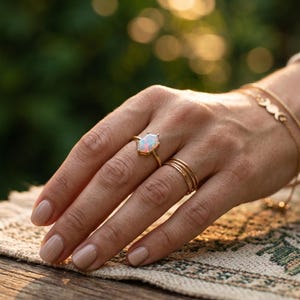 May include: A hand displays a gold ring featuring a hexagonal opal gemstone. Several thin gold rings and a gold bracelet are also visible. The opal has a white and iridescent appearance. The background is blurred.