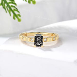 Pode incluir: Um anel de ouro com uma pedra preta retangular, com delicadas inclusões brancas. A banda é adornada com pequenos detalhes quadrados e minúsculas pedras transparentes, criando uma peça de joalheria clássica e elegante.