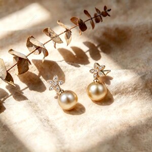 Könnte beinhalten: Ein Paar goldfarbene Ohrhänger mit einer kugelförmigen, perlenartigen Kugel, die an einem blumenförmigen Stecker hängt. Der Blumenstecker ist mit kleinen, klaren, funkelnden Steinen besetzt, die in einen metallischen Rahmen gefasst sind.