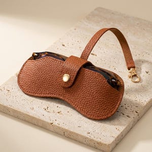 May include: A brown leather glasses case with a matching strap and gold-coloured clasp. The case is closed with a snap and holds a pair of tortoiseshell glasses. The case has a textured surface.