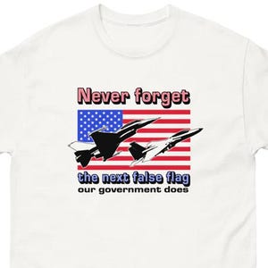 May include: White t-shirt with a graphic design featuring a stylized American flag with fighter jets and the text "Never forget the next false flag our government does."