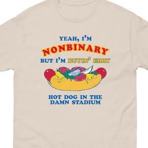 May include: Beige t-shirt with a graphic of a hot dog and text. The text reads "YEAH, I'M NONBINARY BUT I'M BUYIN' ERRY HOT DOG IN THE DAMN STADIUM" in blue, red, and yellow. The hot dog graphic has red, green, and yellow accents.