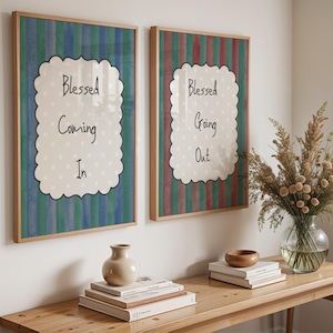 May include: Two framed art prints with wooden frames hang on a white wall. The prints feature a scalloped white shape with the words "Blessed Coming In" and "Blessed Going Out" on a striped background.