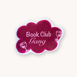 Könnte beinhalten: Ein wolkenförmiger Aufkleber mit dunkelmagenta Hintergrund und dem weißen Text "Book Club Gang" in der Mitte. Zwei hellrosa Weinglas-Illustrationen befinden sich links und rechts vom Text innerhalb der Wolkenform auf dem Aufkleber.