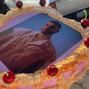 May include: A rectangular cake with a photo of a person on top, decorated with pink and yellow frosting and topped with cherries. The cake has a light pink layer and is surrounded by a border of yellow frosting.