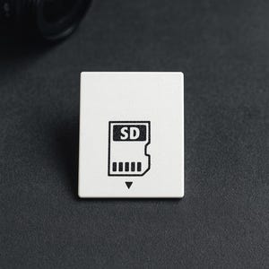 Puede incluir: Adaptador de tarjeta SD blanco con un símbolo de tarjeta SD negro. El adaptador cuadrado está sobre una superficie oscura. Una lente de cámara negra es parcialmente visible en el fondo.