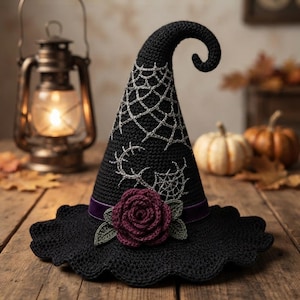 Puede incluir: Un sombrero de bruja negro de ganchillo con punta curvada, decorado con telarañas plateadas bordadas, una cinta de terciopelo morado y una rosa de ganchillo de color rojo oscuro con hojas verdes en el ala ancha y festoneada.