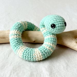 Può includere: Un giocattolo a forma di anello a forma di serpente all'uncinetto. Il serpente è principalmente azzurro con strisce bianche e ha occhi neri a bottone. Il giocattolo è appoggiato su un pezzo di legno.