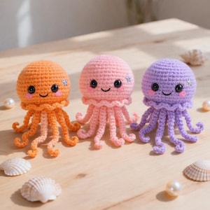 May include: Three handmade crochet jellyfish in orange, pink, and purple. Each has a round head, smiling face, and dangling tentacles. Decorative star accents are on the heads. Seashells and pearls are scattered around the jellyfish.