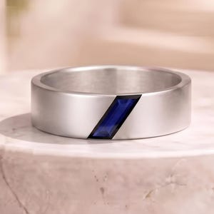 Puede incluir: Un anillo de banda de metal plateado que presenta una única piedra preciosa de color azul intenso, con corte rectangular inclinado y engastada al ras de la superficie. El anillo tiene un acabado mate liso y un perfil plano de diseño geométrico.