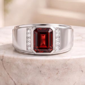 Pode incluir: Um anel de tom prateado com uma grande pedra preciosa retangular de cor vermelho-escura em corte esmeralda no centro. A pedra é ladeada em ambos os lados por uma fila vertical de pequenas pedras de destaque redondas e claras no aro.