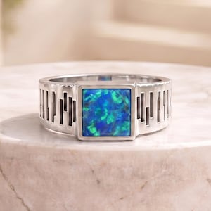 May include: A silver-toned ring featuring a square-cut, iridescent blue opal inlay. The band is constructed with a series of vertical, rectangular cutouts that create a repeating geometric pattern around the entire circumference of the ring.