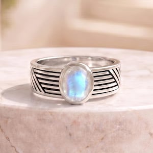 May include: A silver ring featuring a central oval-shaped iridescent moonstone cabochon. The band is crafted from polished silver and displays a series of etched horizontal and diagonal black lines that create a textured geometric pattern around the stone.