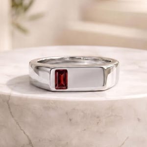 Pode incluir: Um anel de prata com uma pedra preciosa retangular vermelha escura. O anel está sobre uma superfície de mármore branco, mostrando seu metal polido e a pedra preciosa. O design do anel é moderno e minimalista.