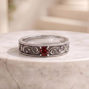 Pode incluir: Um anel de prata com uma pedra preciosa vermelha escura de corte redondo ao centro. A superfície metálica é gravada com padrões de redemoinhos, luas crescentes e pequenas estrelas, contornada por uma textura delicada de contas nas bordas.