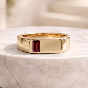 Op de afbeelding: Een gouden ring met een rechthoekige rode edelsteen in het midden. De ring wordt gepresenteerd op een wit marmeren oppervlak, wat de gepolijste afwerking en het elegante ontwerp benadrukt. De ring is ongeveer 1,5 cm breed.