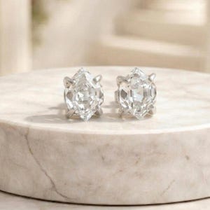 Könnte beinhalten: Ein Paar Ohrstecker mit Diamanten im Marquise-Schliff. Die Ohrringe sind in einem silberfarbenen Metall gefasst und werden auf einer weißen Marmoroberfläche präsentiert. Die Ohrringe sind der Fokus des Bildes.