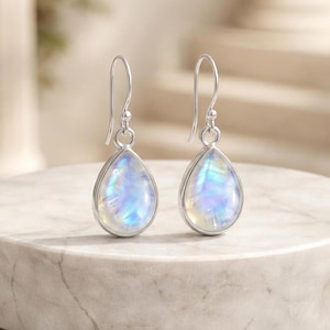 Pode incluir: Um par de brincos em forma de gota com armação prateada. Os brincos apresentam uma pedra iridescente perolada que reflete tons de azul, branco e amarelo. São exibidos numa superfície de mármore.