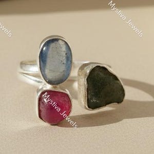 May include: A silver ring featuring three gemstones: a light blue oval, a deep red square, and a dark green, irregularly shaped stone. The ring is set against a neutral background, highlighting the colors and textures of the stones.