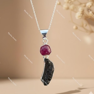 May include: A silver necklace with a pendant featuring a dark red gemstone and a dark, irregular-shaped stone. The necklace has a delicate chain and a triangular silver element. The background is a soft, neutral color.