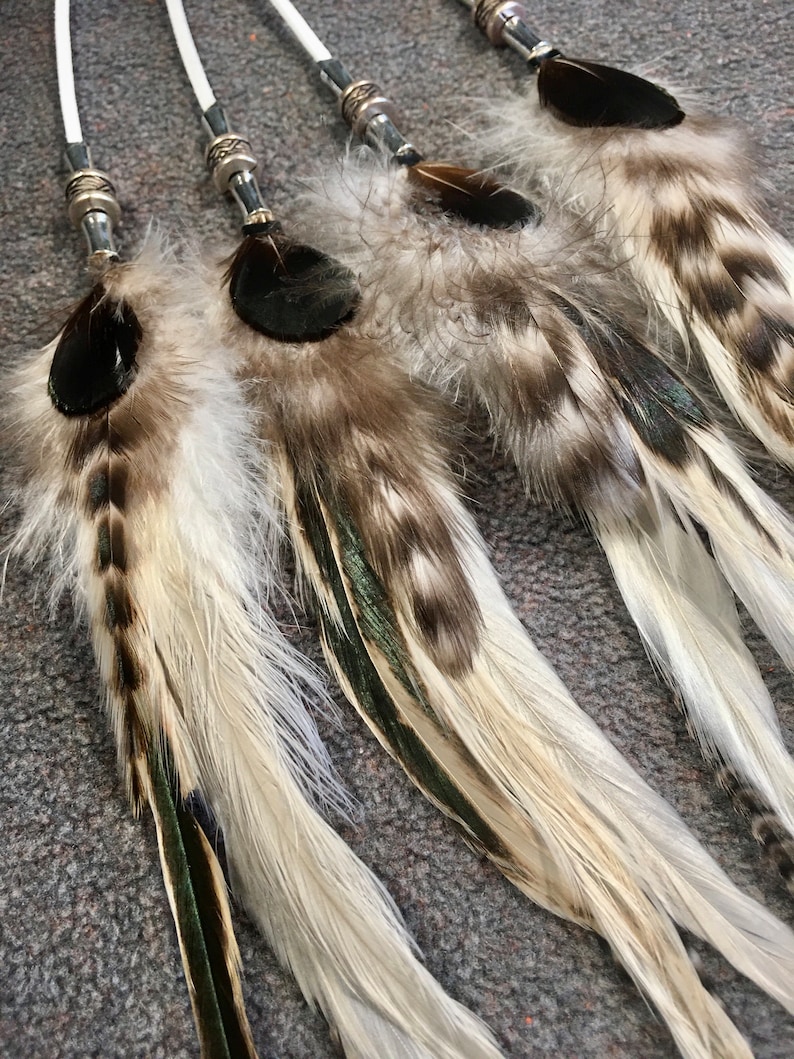 May include: Five white and brown feathers with black tips and green accents are tied together with white string and silver beads.